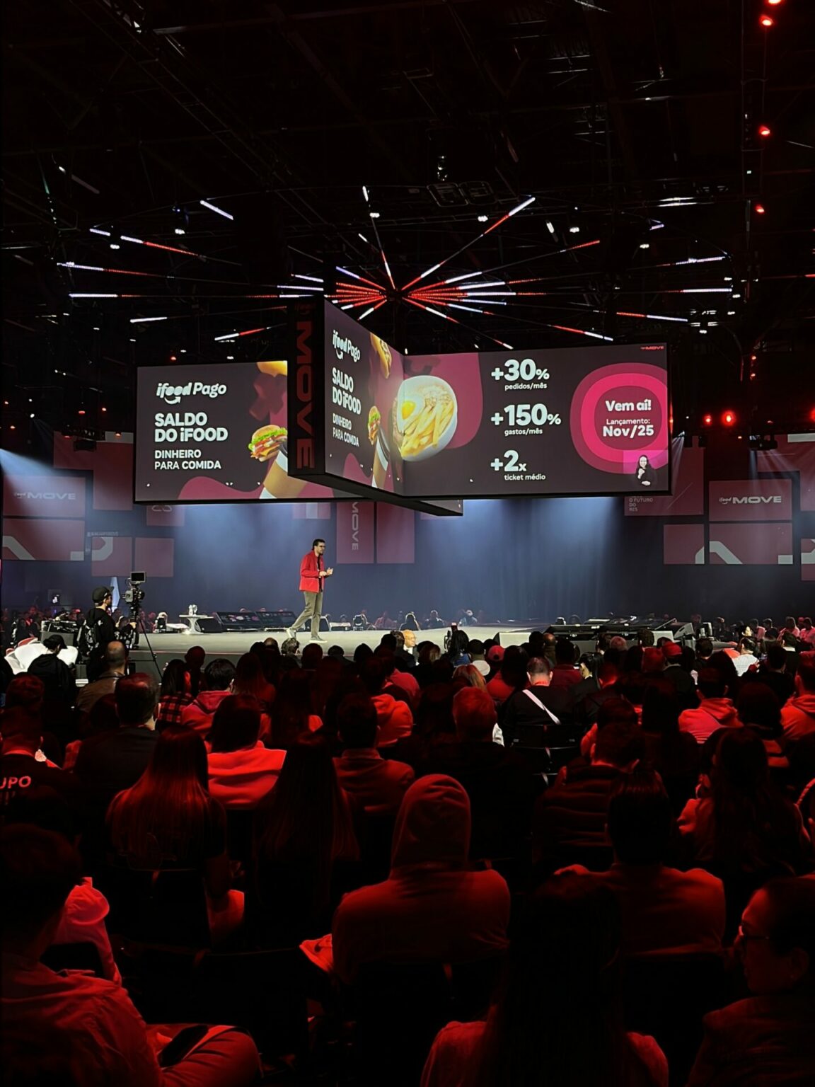 Evento do iFood Move 2025 em palco com telões iluminados e plateia lotada em ambiente de conferência, com luzes cênicas vermelhas e destaque para apresentações no centro.